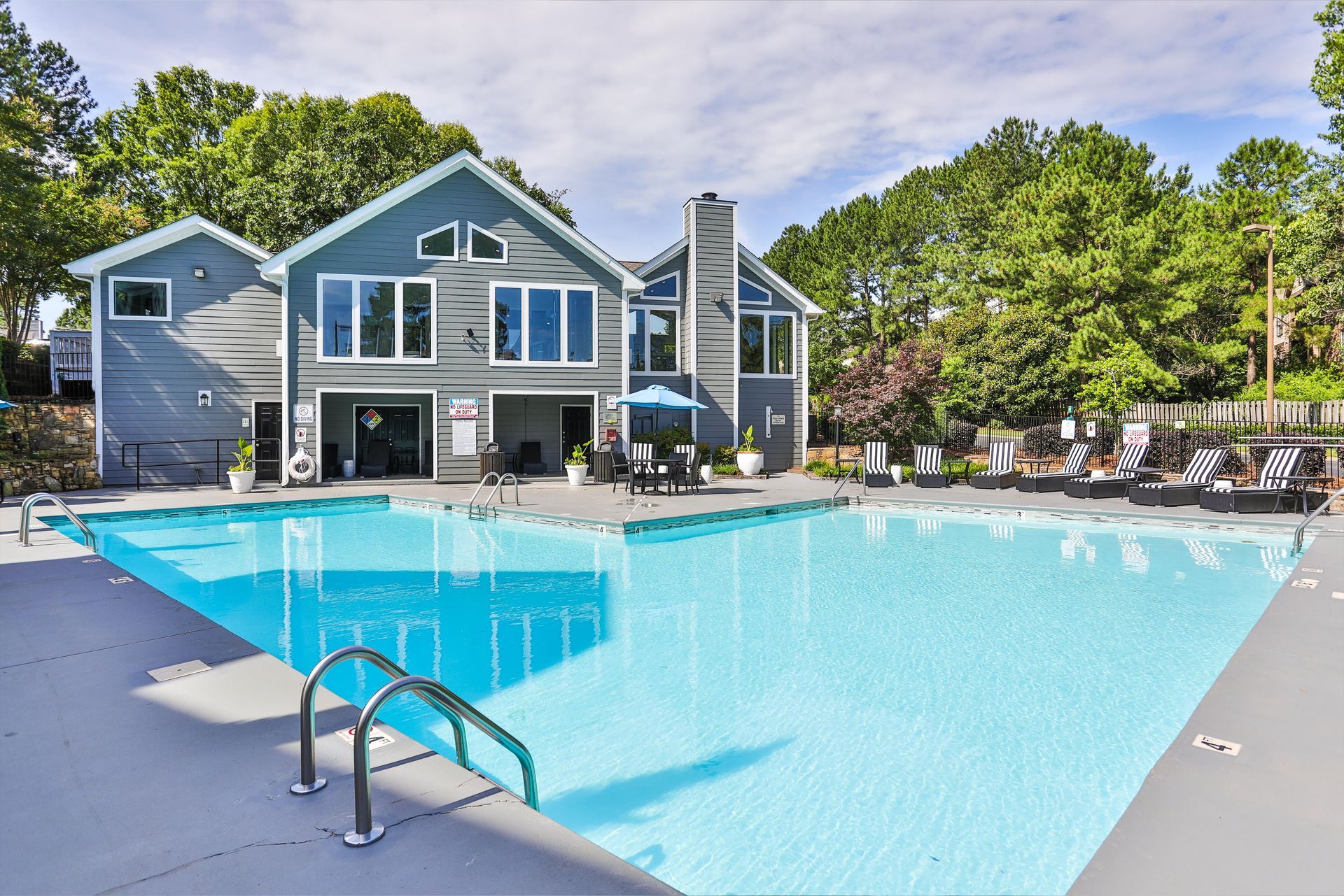 Swimming pool in front of a blue-gray building with a patio, trees, and lounge chairs on the side. Our apartments in Charlotte, NC offer a convenient location and top-notch amenities. Call today to schedule your tour!
