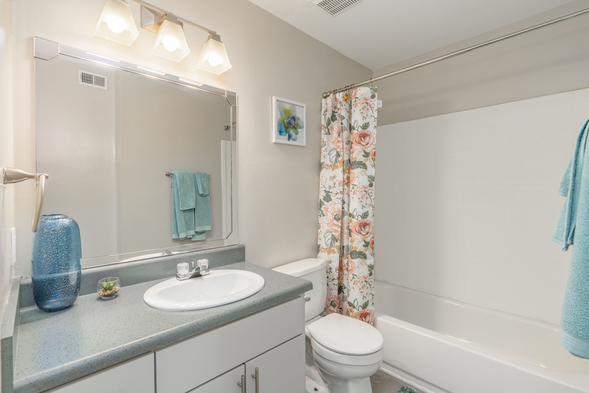Bathroom with a white toilet, sink, and bathtub. Floral shower curtain and turquoise towels. Contact us today to take a tour of our 1, 2, and 3-bedroom apartments for rent in Charlotte, NC.