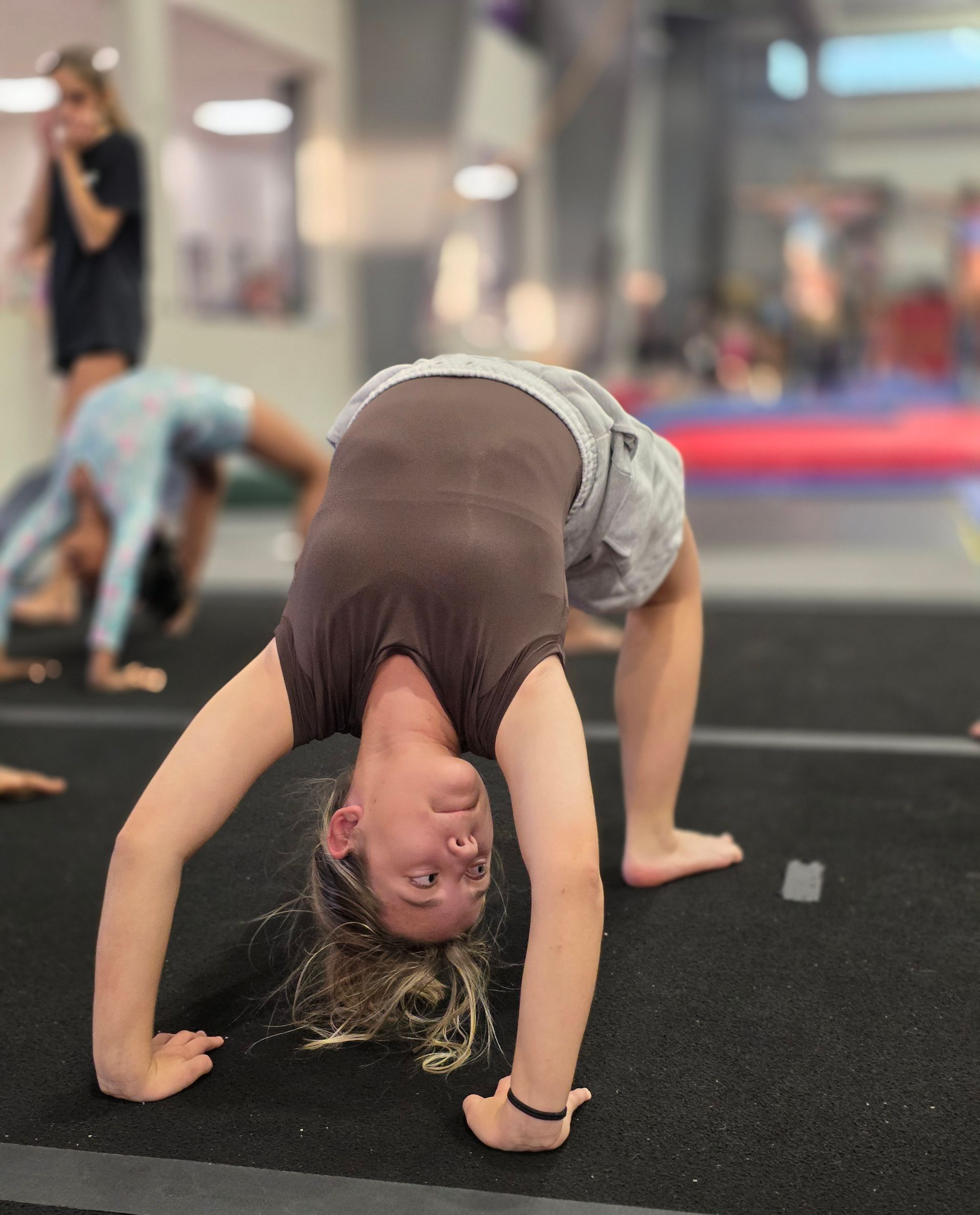 A person in a brown top and gray shorts performs a backbend on a black gym mat with others practicing in the background.