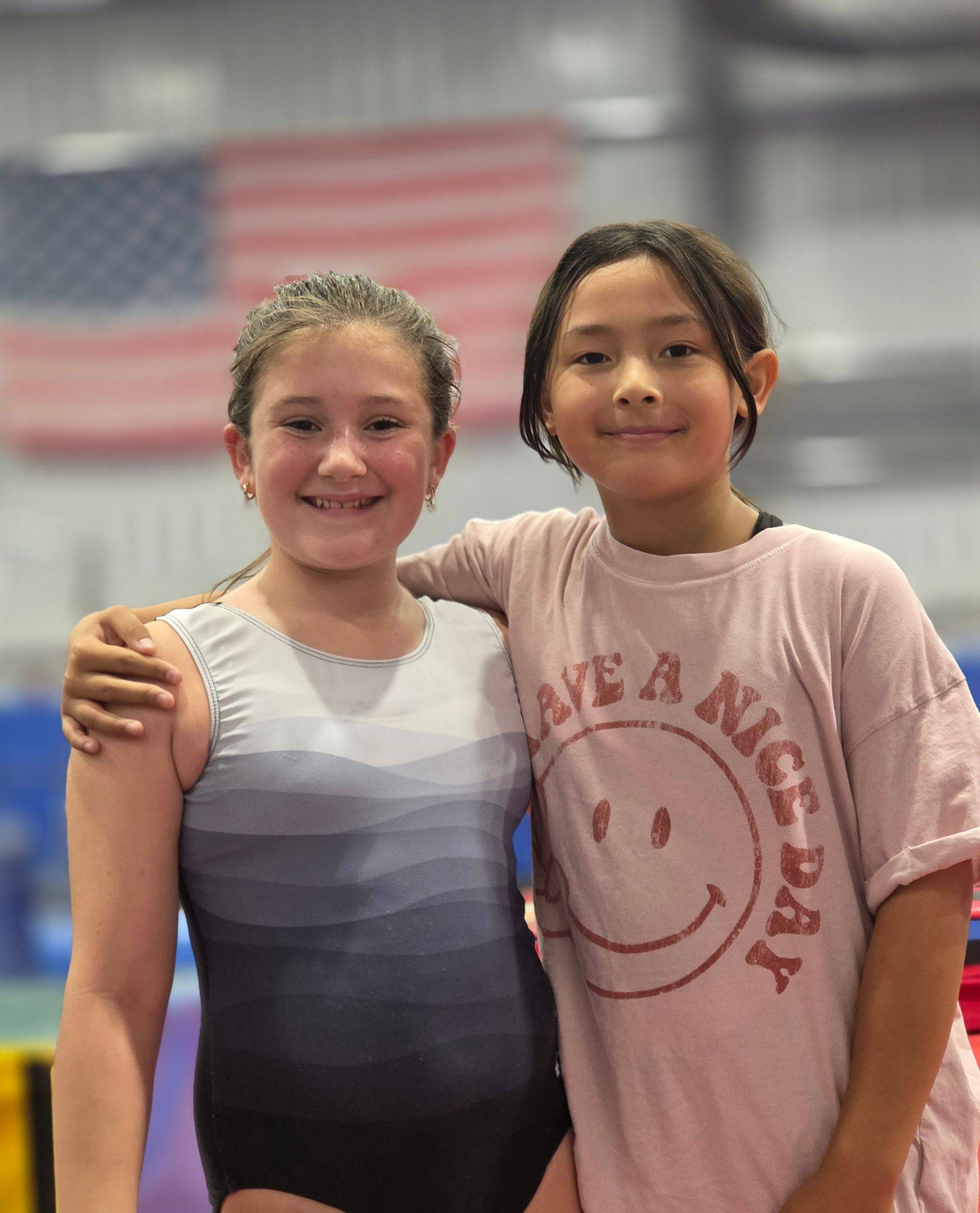 Two children smiling and posing for a photo in a gym with an American flag in the background.