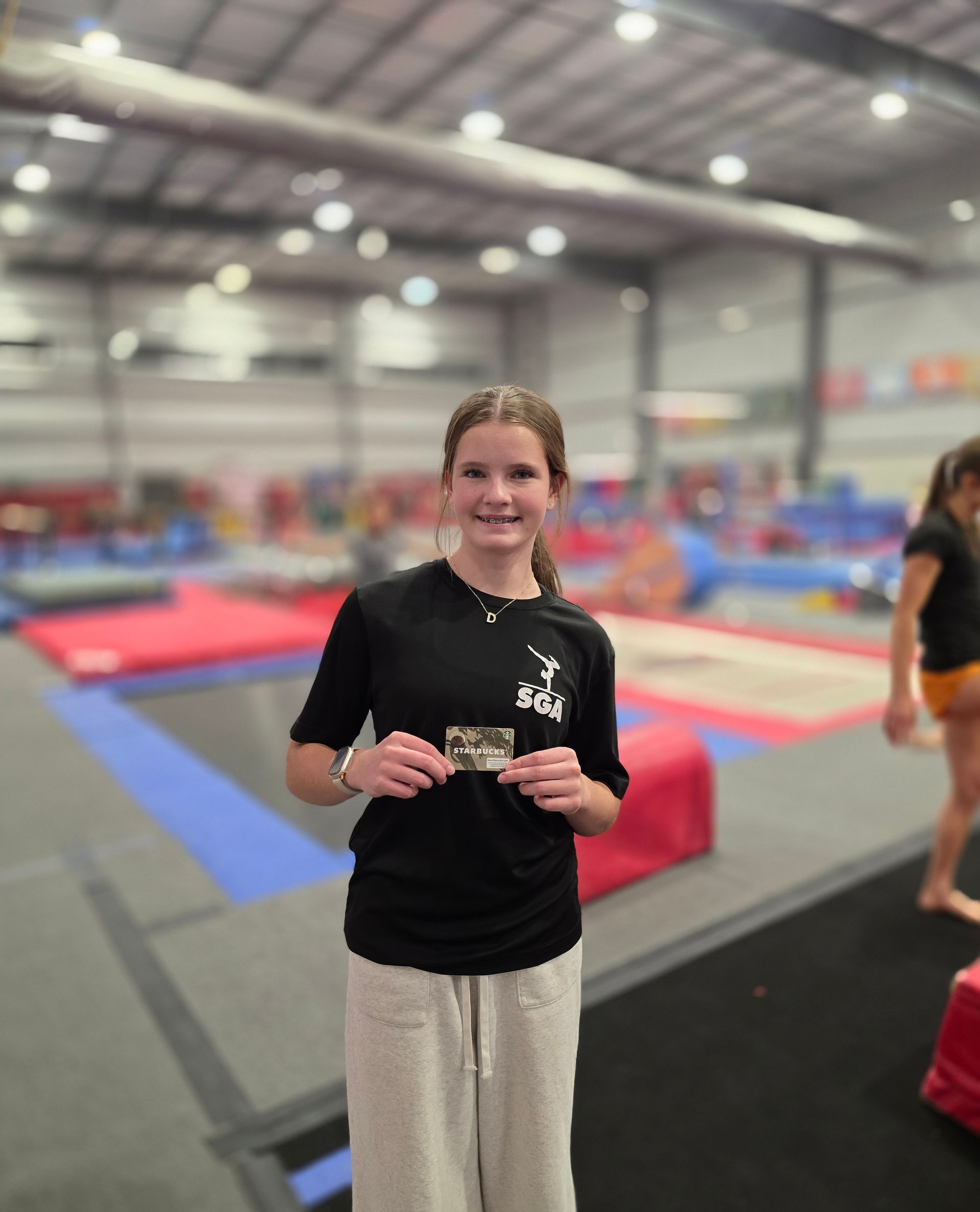 A smiling person stands in a gymnastics gym, holding a small card, wearing a black t-shirt and grey sweatpants.