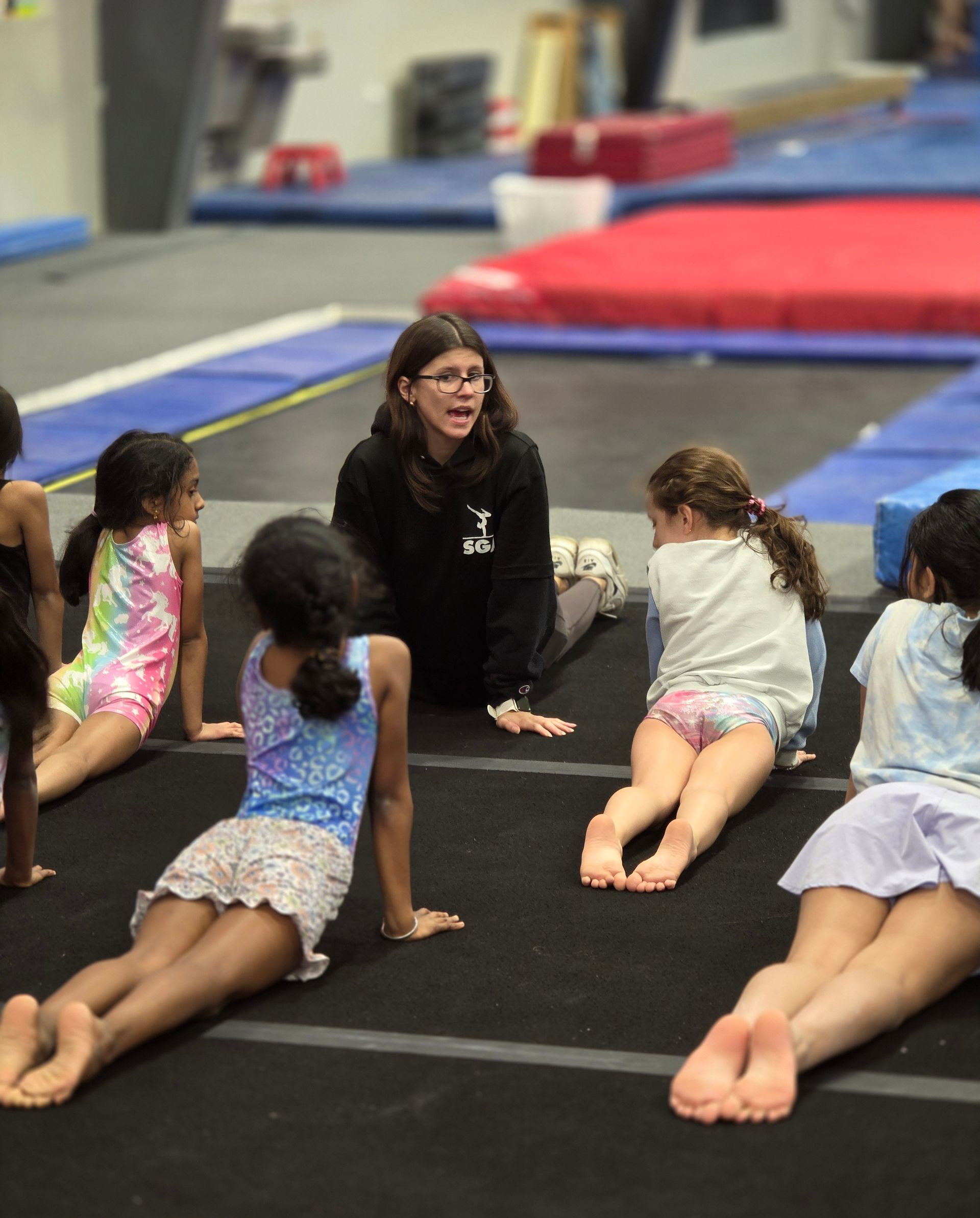 A coach leads a group of students in a cobra yoga stretch on a black floor in a gymnastics training center.