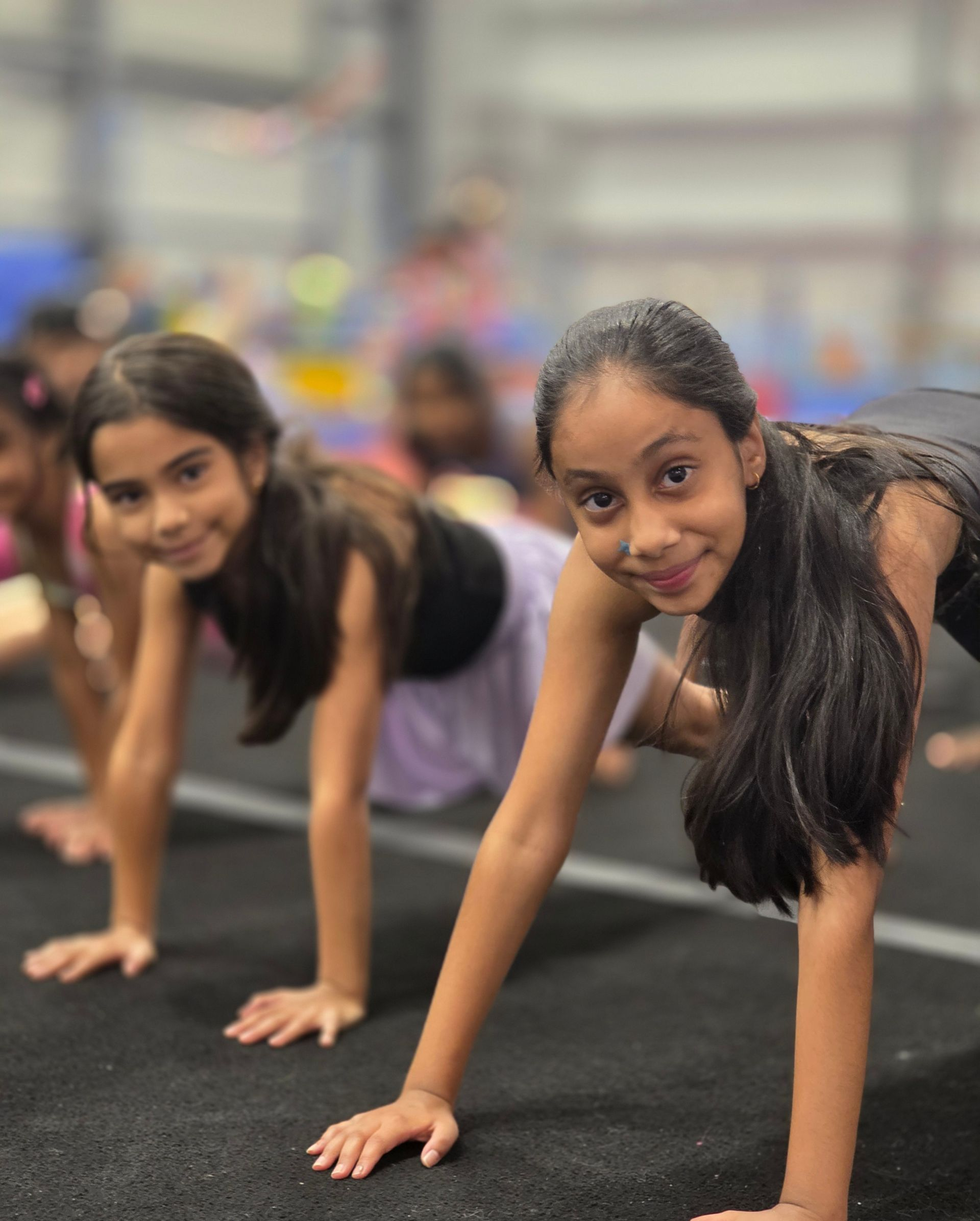 Two people smiling while in a plank position on a black gym mat in a studio.
