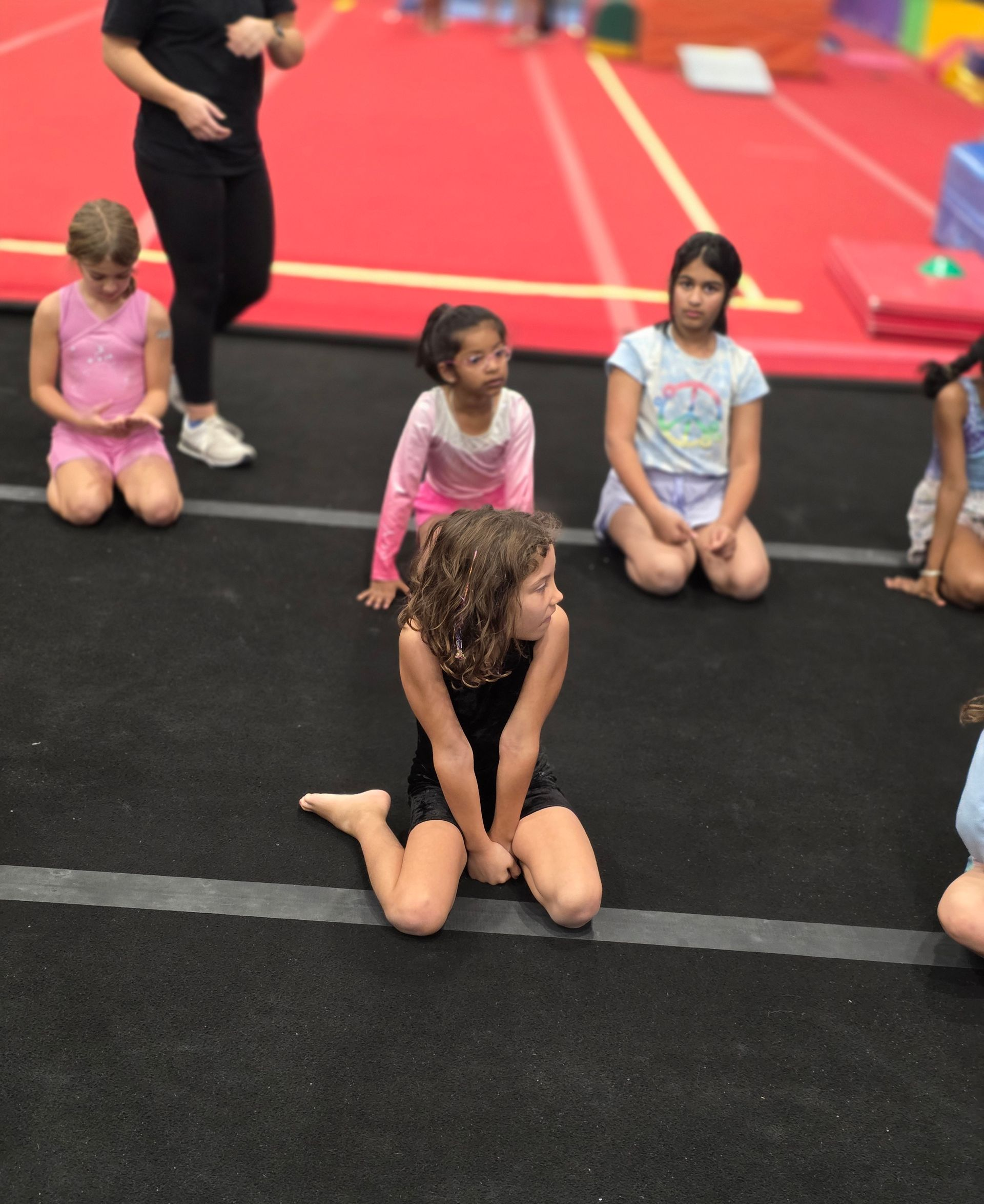 A group of children and an instructor kneel on a gymnastics floor during a class.