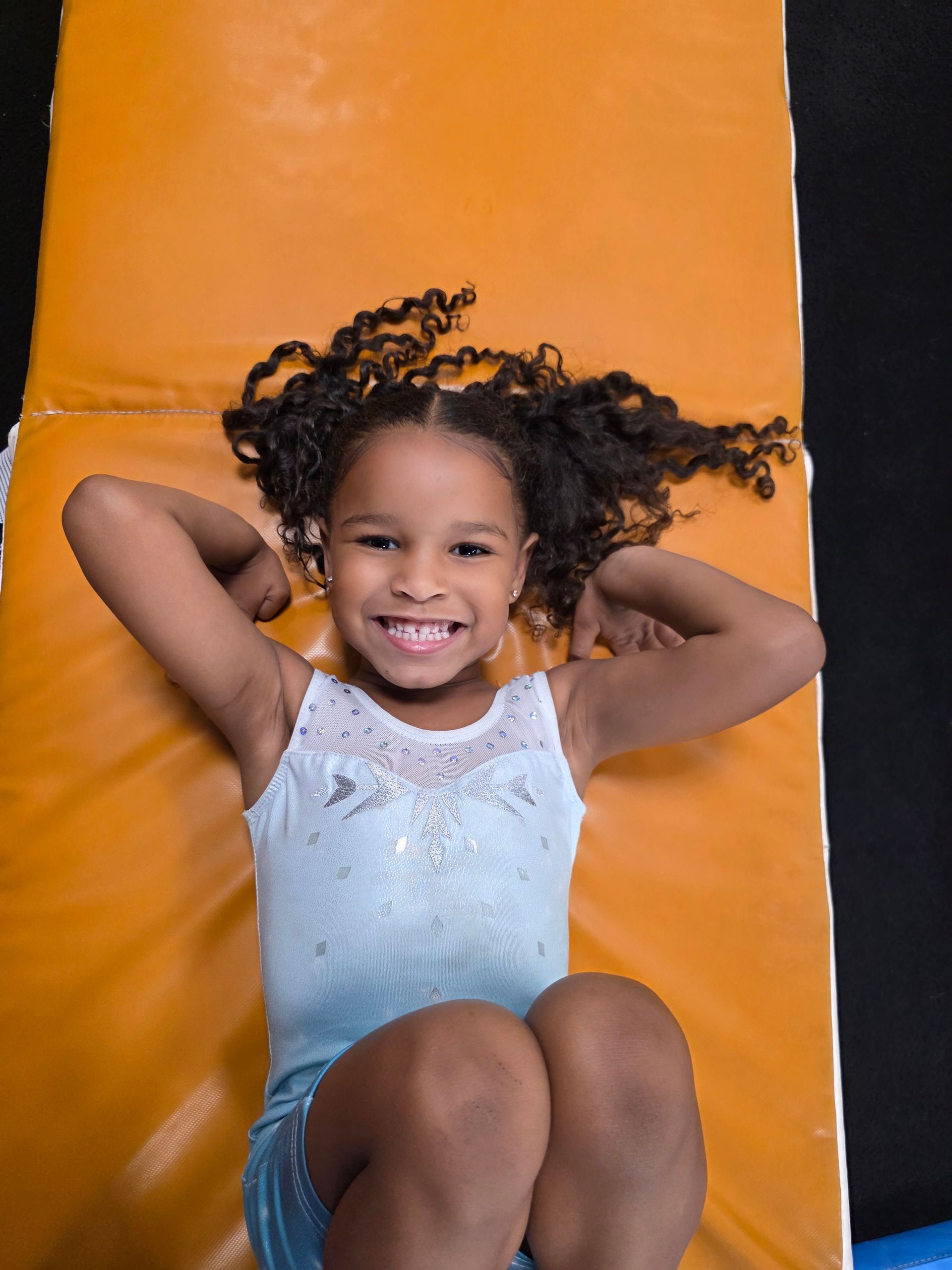 A smiling child in a light blue leotard lies on an orange gym mat with their arms behind their head.