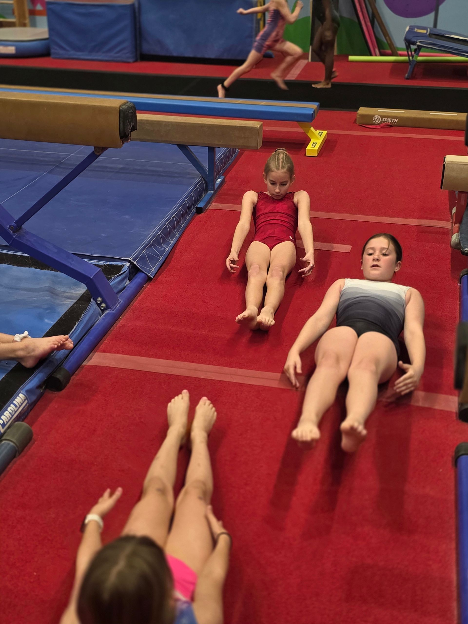 A gymnast in a red leotard performs a back tuck on a trampoline in a gym.