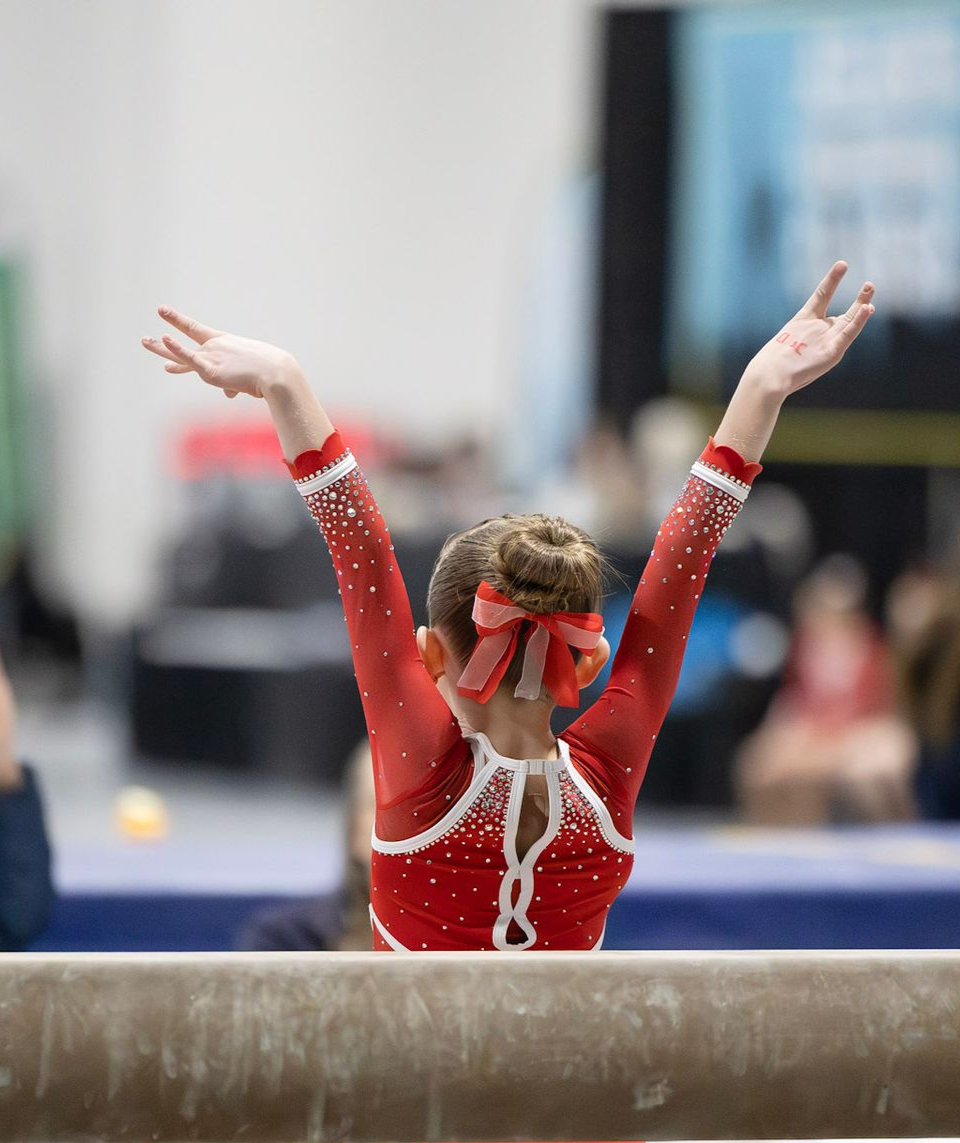 A gymnast in a red leotard with a matching bow stands on a balance beam with arms raised in a pose during a competition.
