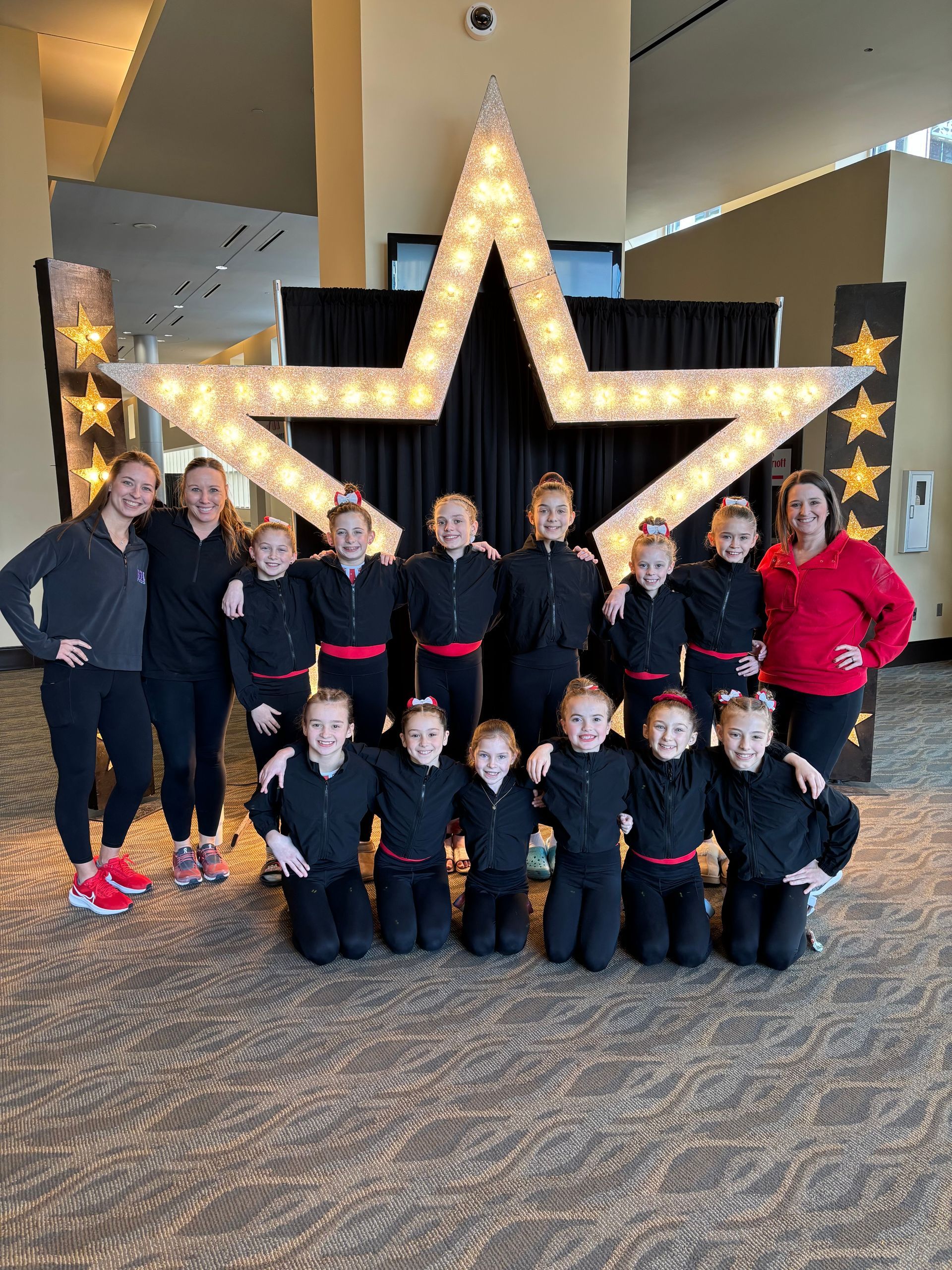 A group of dancers in black uniforms poses with two coaches in front of a large, illuminated star backdrop.
