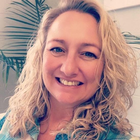 Smiling woman with curly blonde hair in a light blue top, indoors with a plant in the background