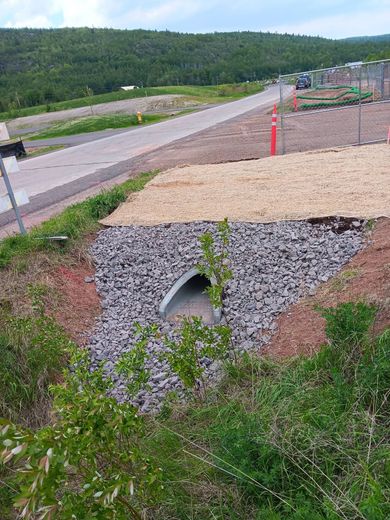 Roadside drainage culvert with rock lining beside a paved lane and grassy embankment under overcast skies
