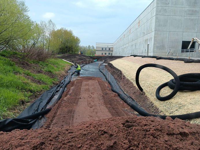 Construction site with a dirt trench, black drainage tubing, and a large building wall beside a grassy slope