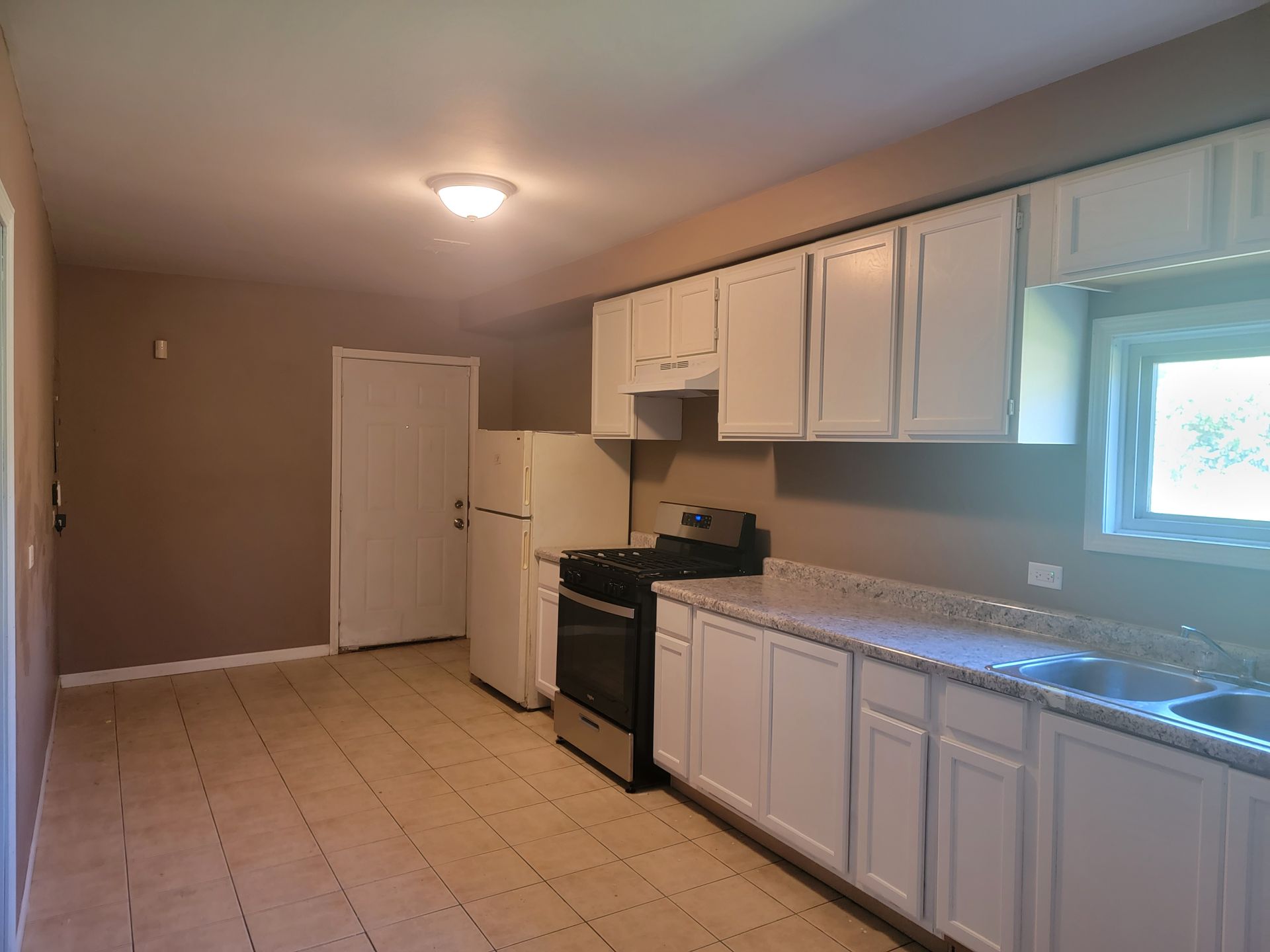 An empty kitchen with white cabinets , a stove , refrigerator , sink and window.
