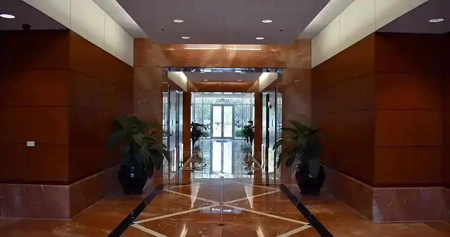 Elegant building lobby with polished marble floors, wood paneling, and potted plants flanking an entrance.