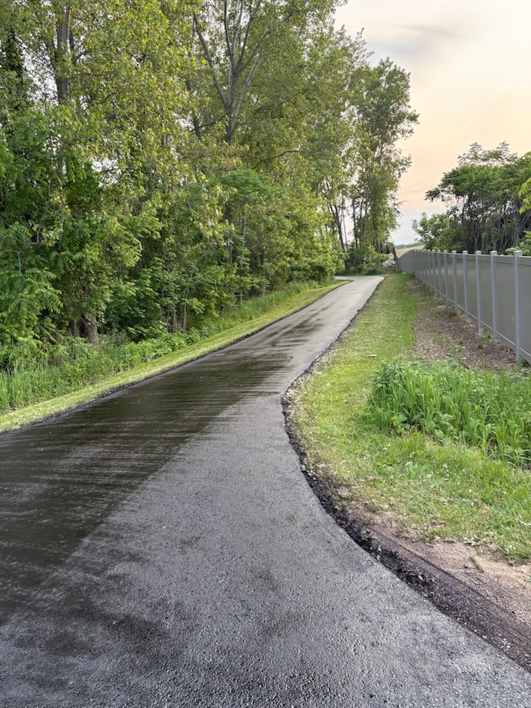 Wet paved path curving through green trees beside a chain-link fence at dusk