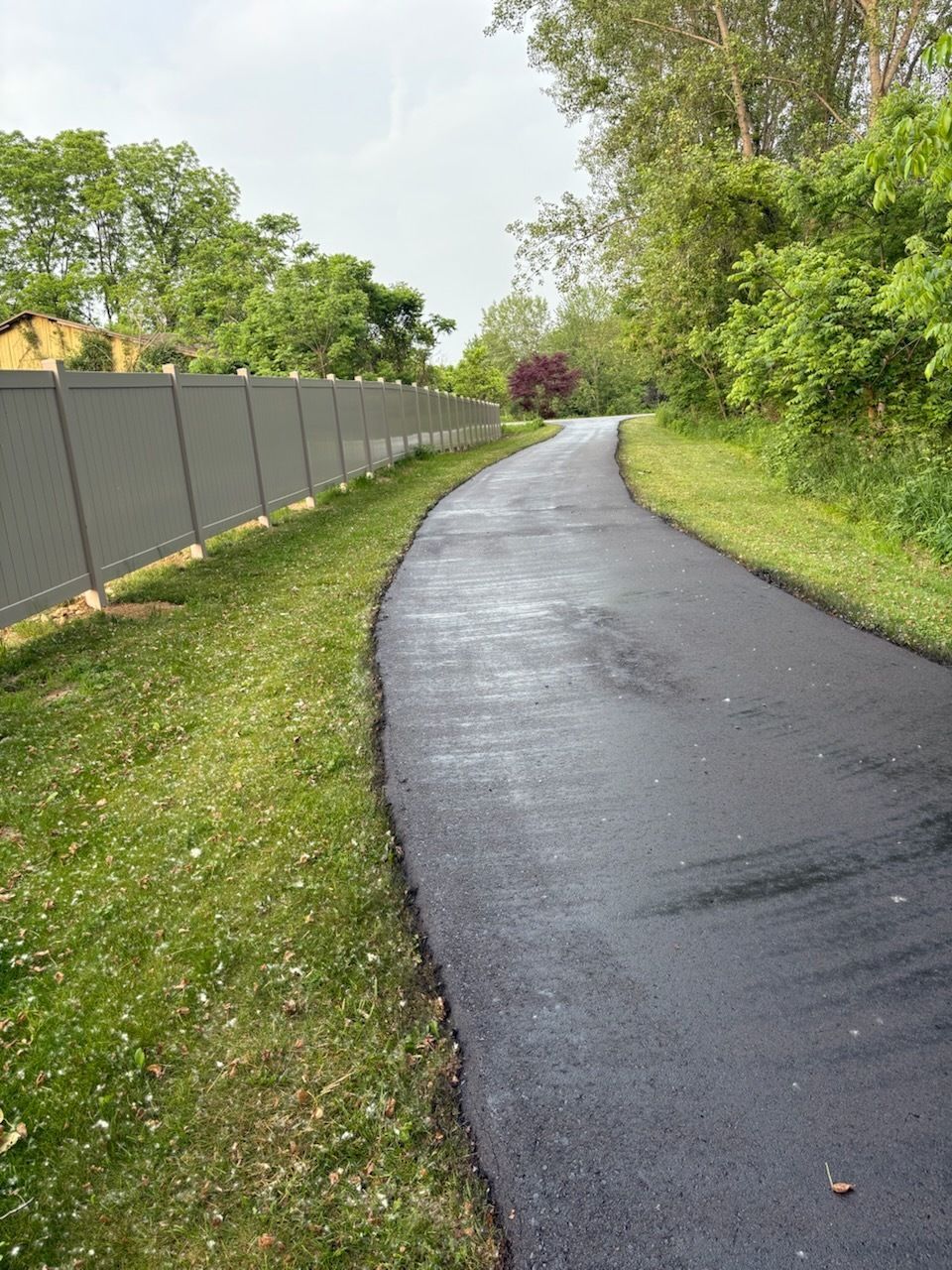 Wet paved path curving through a green park with trees and a fence on the left