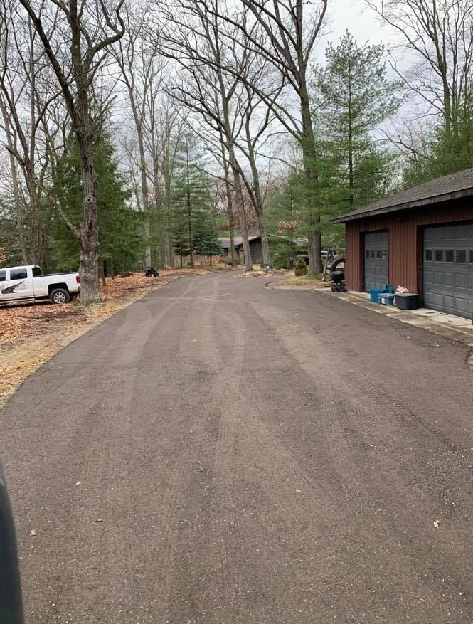 Driveway leading to a wooded residential area beside a brick garage under overcast skies.