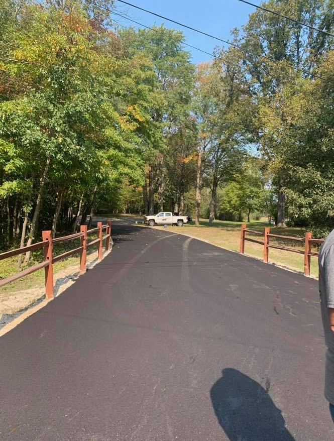 Paved road through a wooded park, lined with wooden rails, with a white car ahead.
