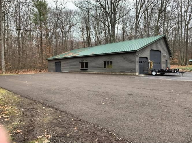 Gray cabin with green roof beside a gravel lot in a leafless wooded area