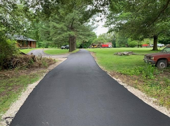 Fresh blacktop driveway through a green yard with trees, a cabin, and parked vehicles in the distance