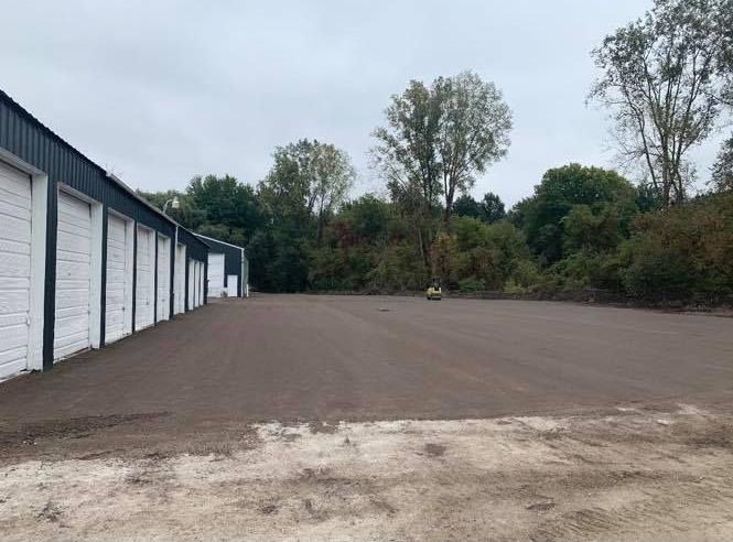 Empty gravel lot beside a row of white garage units and trees under an overcast sky