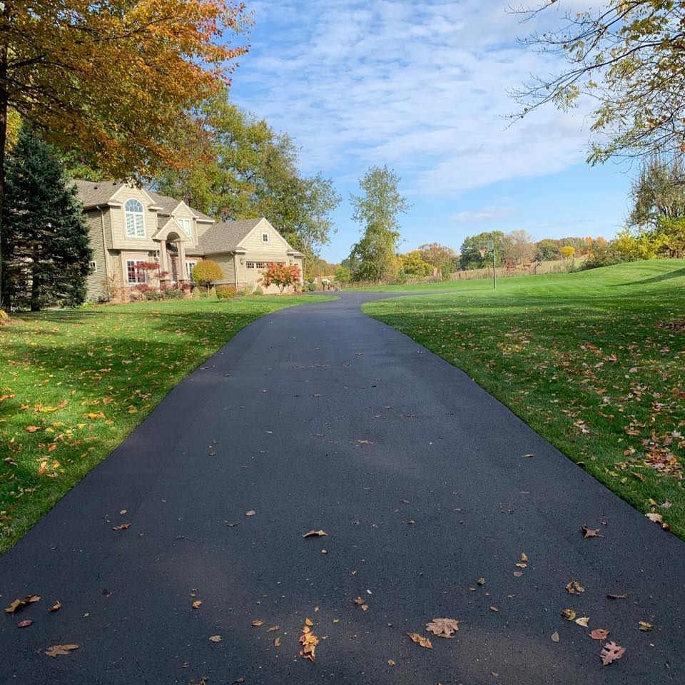 Curved paved driveway beside beige houses, green lawn, and autumn trees under a blue sky.