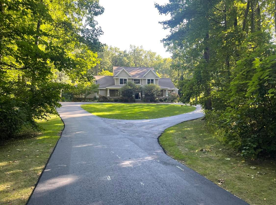 Curved driveway leading to a house surrounded by green trees and lawn in soft sunlight
