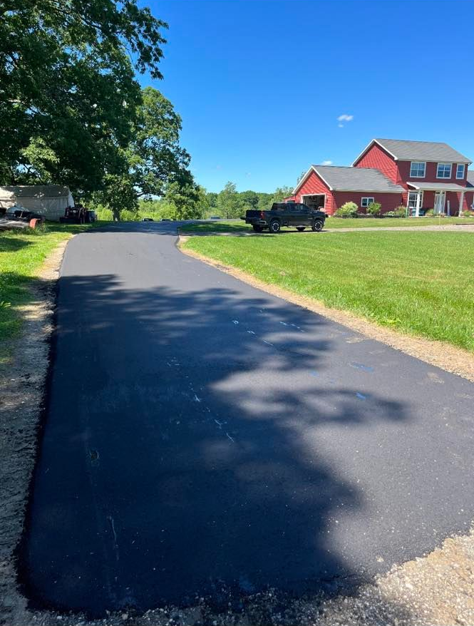 Freshly paved black driveway leading to a red house on a sunny rural property