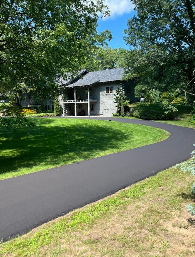 Curving paved path through a green yard leading to a gray house under leafy trees.