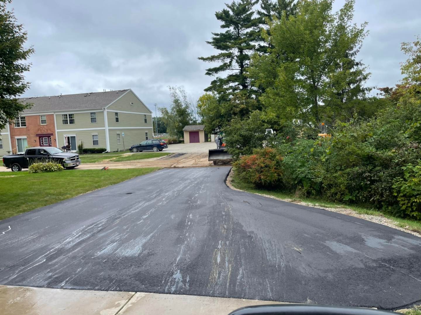 Freshly paved driveway leading to apartment buildings, with trees, shrubs, and parked vehicles on a cloudy day