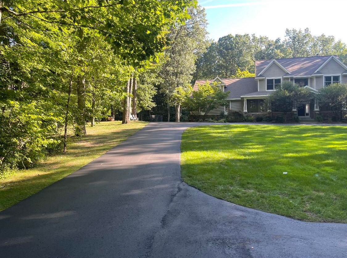 Paved driveway curving past green lawn and trees toward a house in the distance