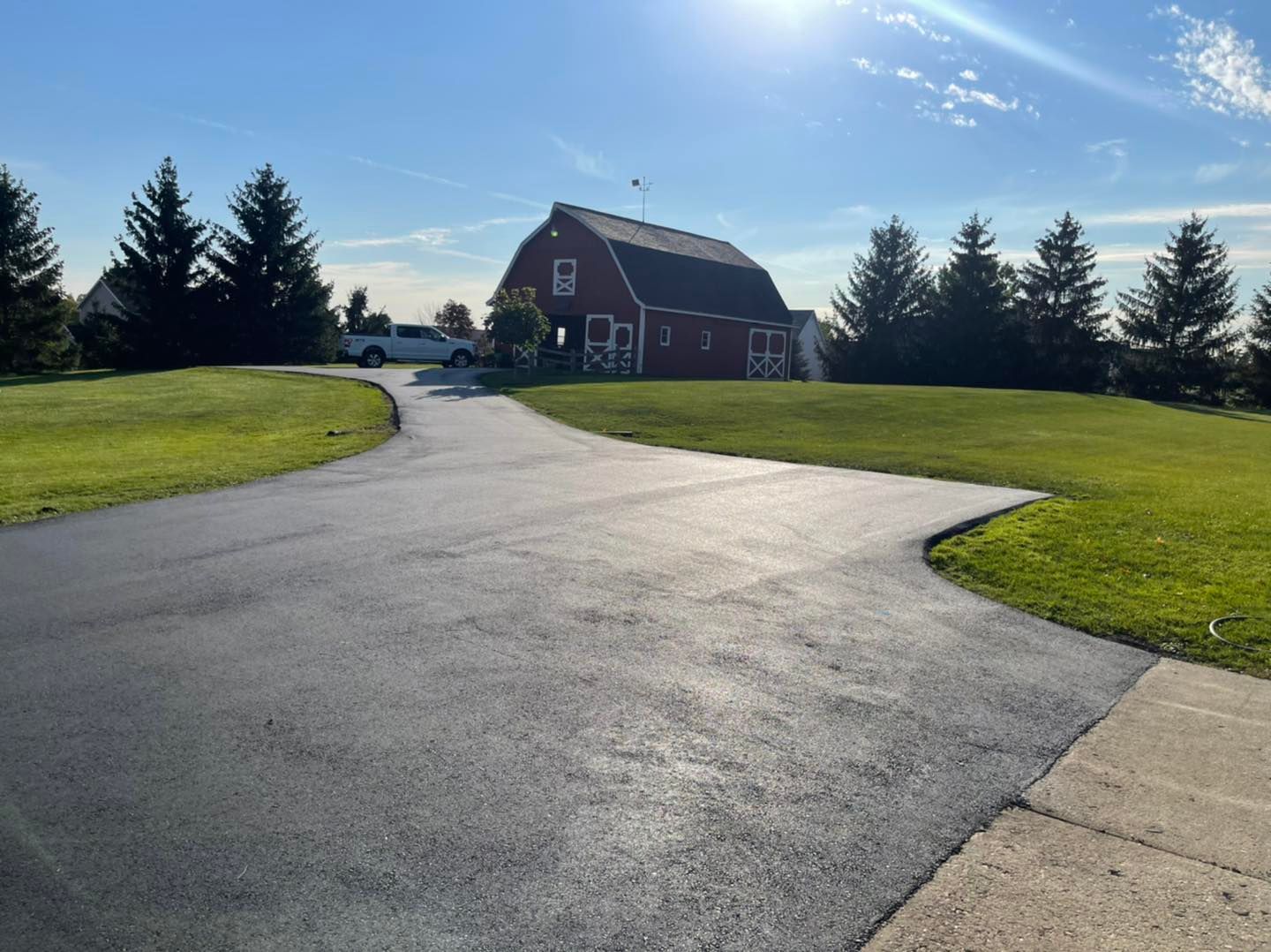 Curved driveway leading to a red barn-style house on a sunny, grassy property with trees.