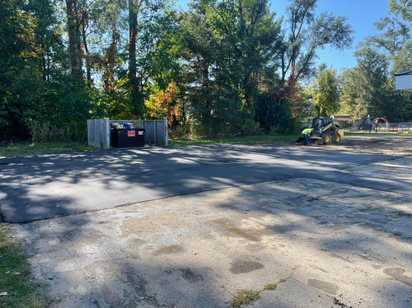 Gravel lot beside trees, with a dumpster and a small excavator in bright sunlight.