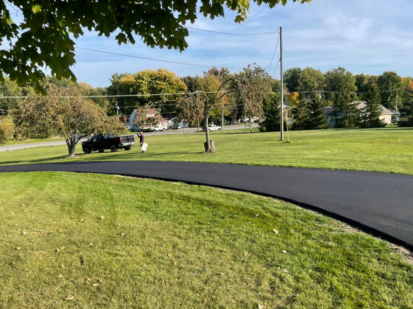 Curved black paved path through a grassy park with trees, a parked car, and a utility pole under a blue sky