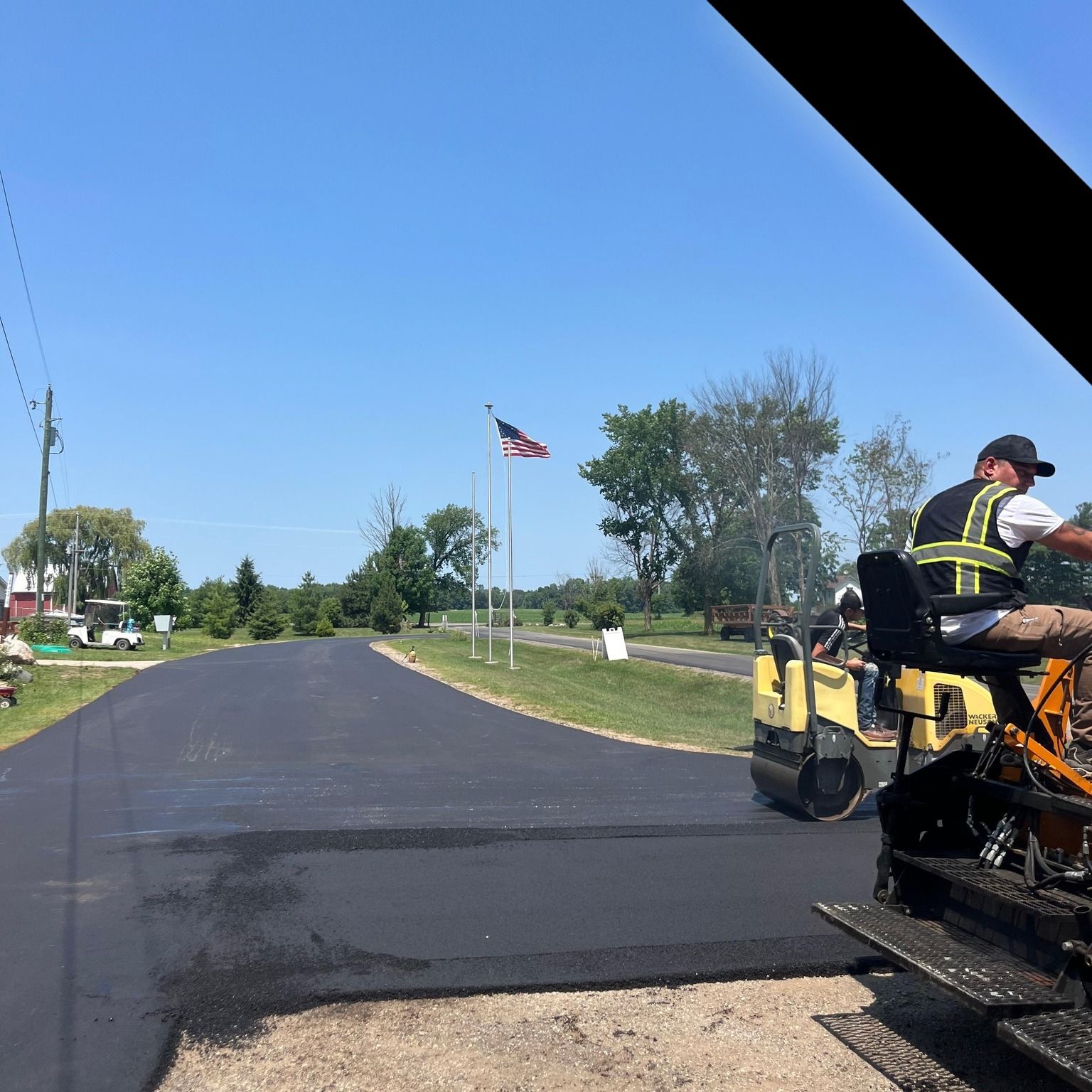 Freshly paved road with a road crew machine on the right and an American flag in the distance.