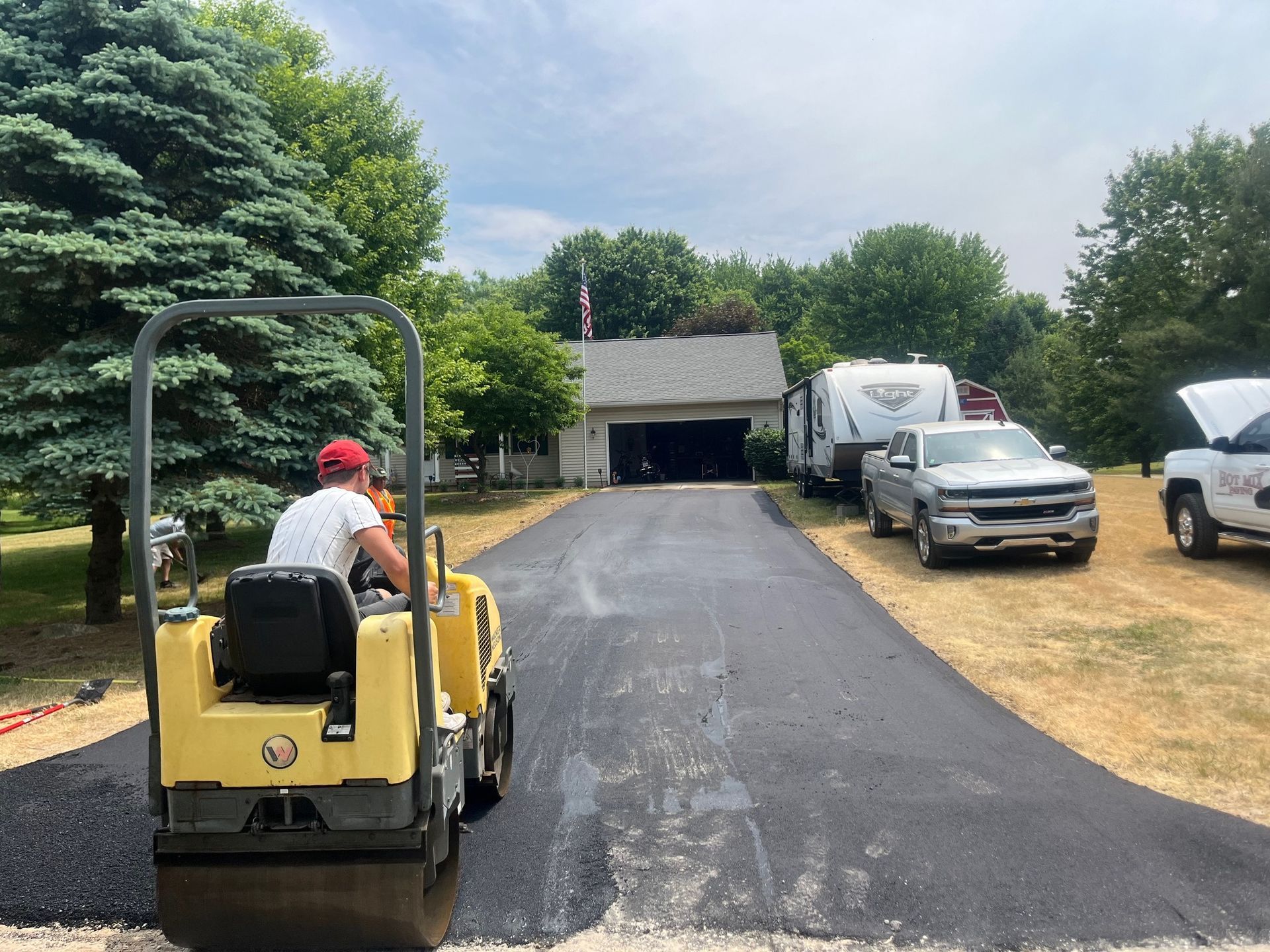Person driving a yellow tractor on a freshly paved driveway toward a garage with parked vehicles nearby