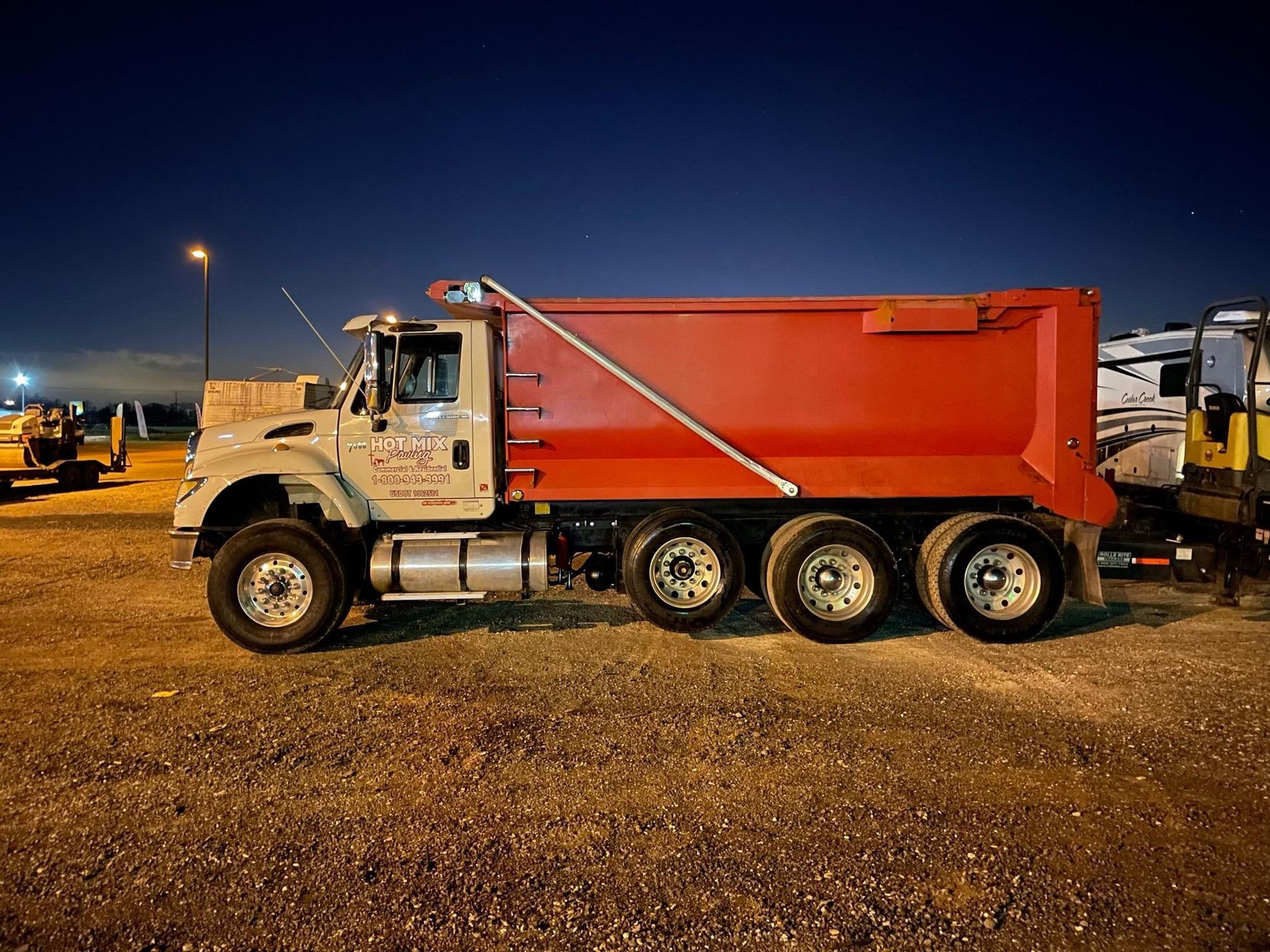 White dump truck with a red bed parked at night on a gravel lot.
