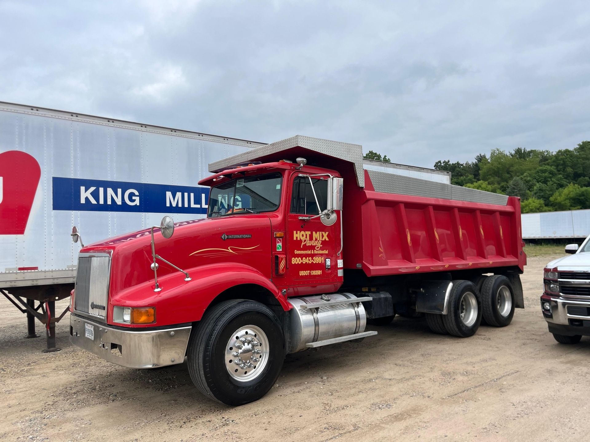 Red dump truck parked on gravel near a King Mill trailer under cloudy skies