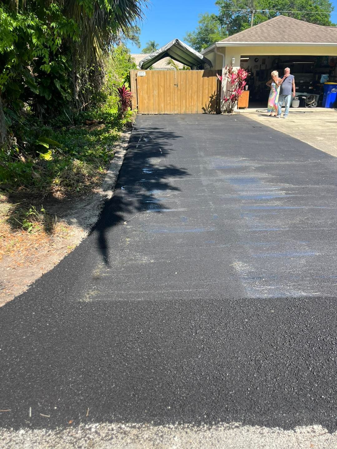 Freshly paved black driveway leading to a wooden fence and garage with people standing nearby