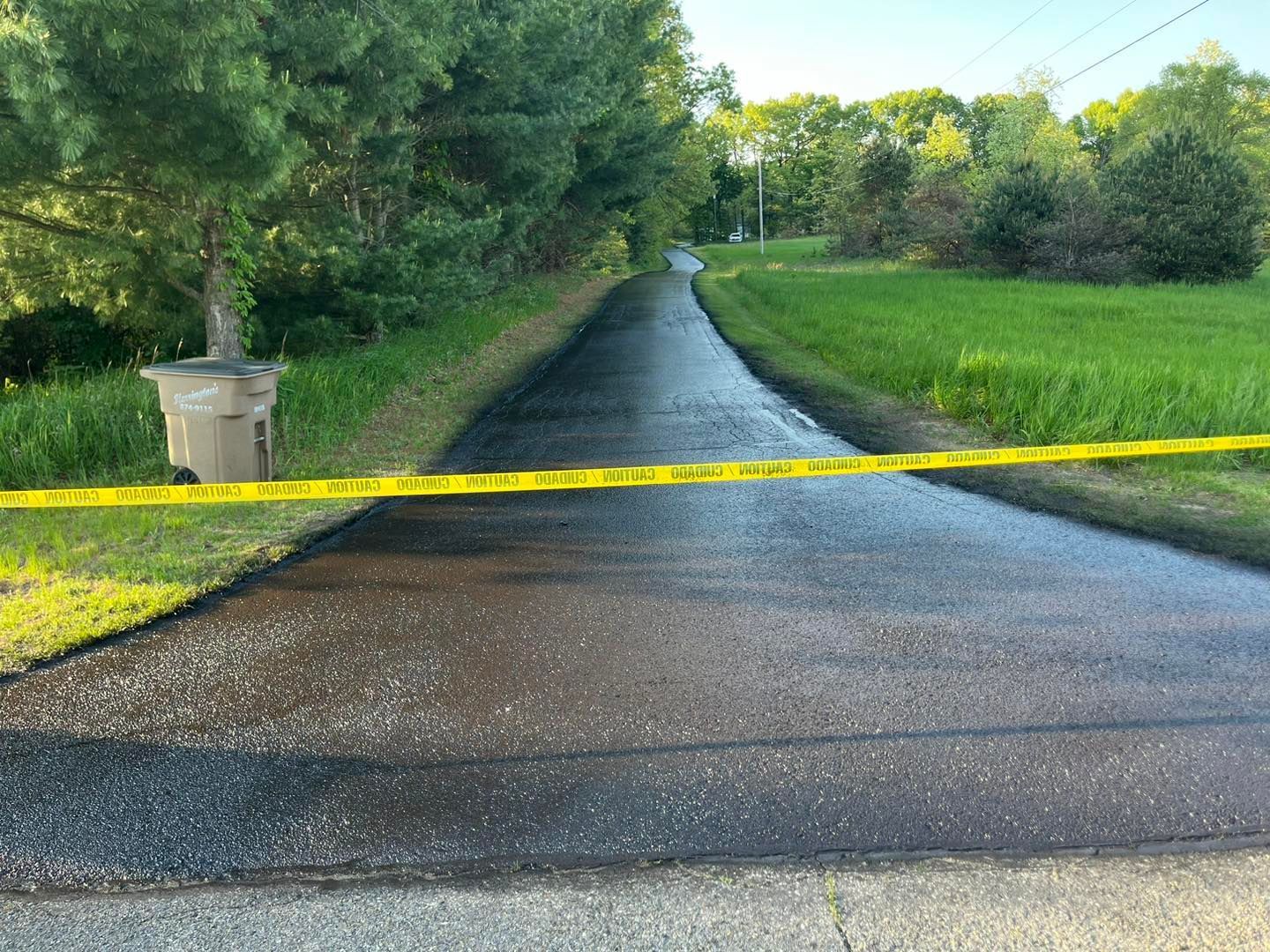Yellow caution tape blocks a wet driveway leading to a tree-lined road after rainfall