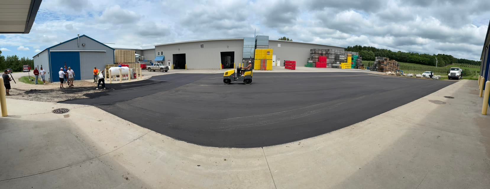 Industrial yard with a wide black asphalt area, buildings, vehicles, and workers under cloudy sky