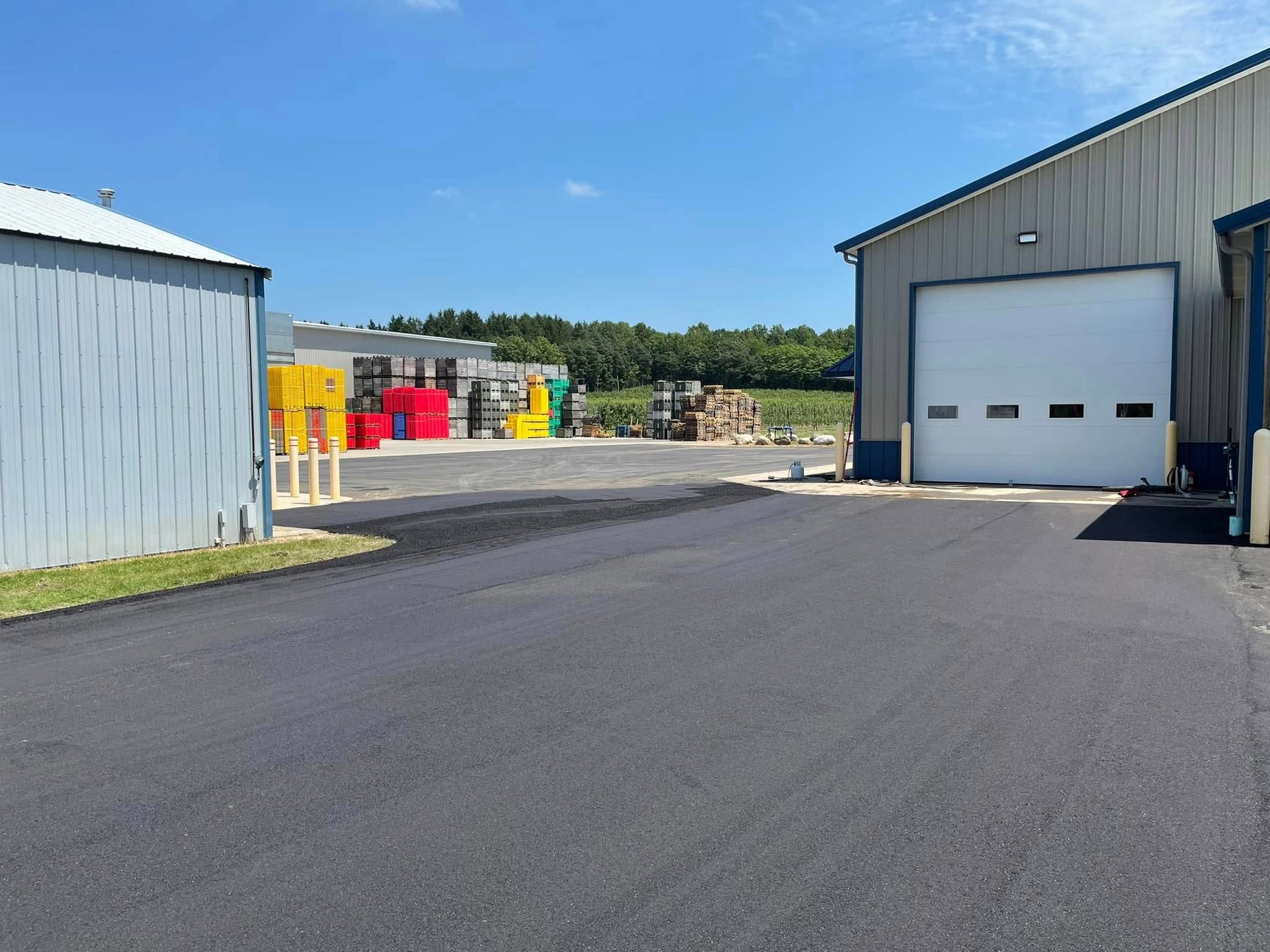 Industrial yard with a large black paved lot, metal buildings, and colorful equipment in the distance
