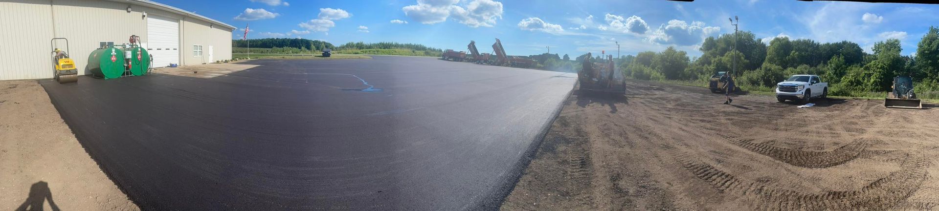 Wide paved parking lot by industrial buildings under a blue sky, with cars and green bins along the left side.