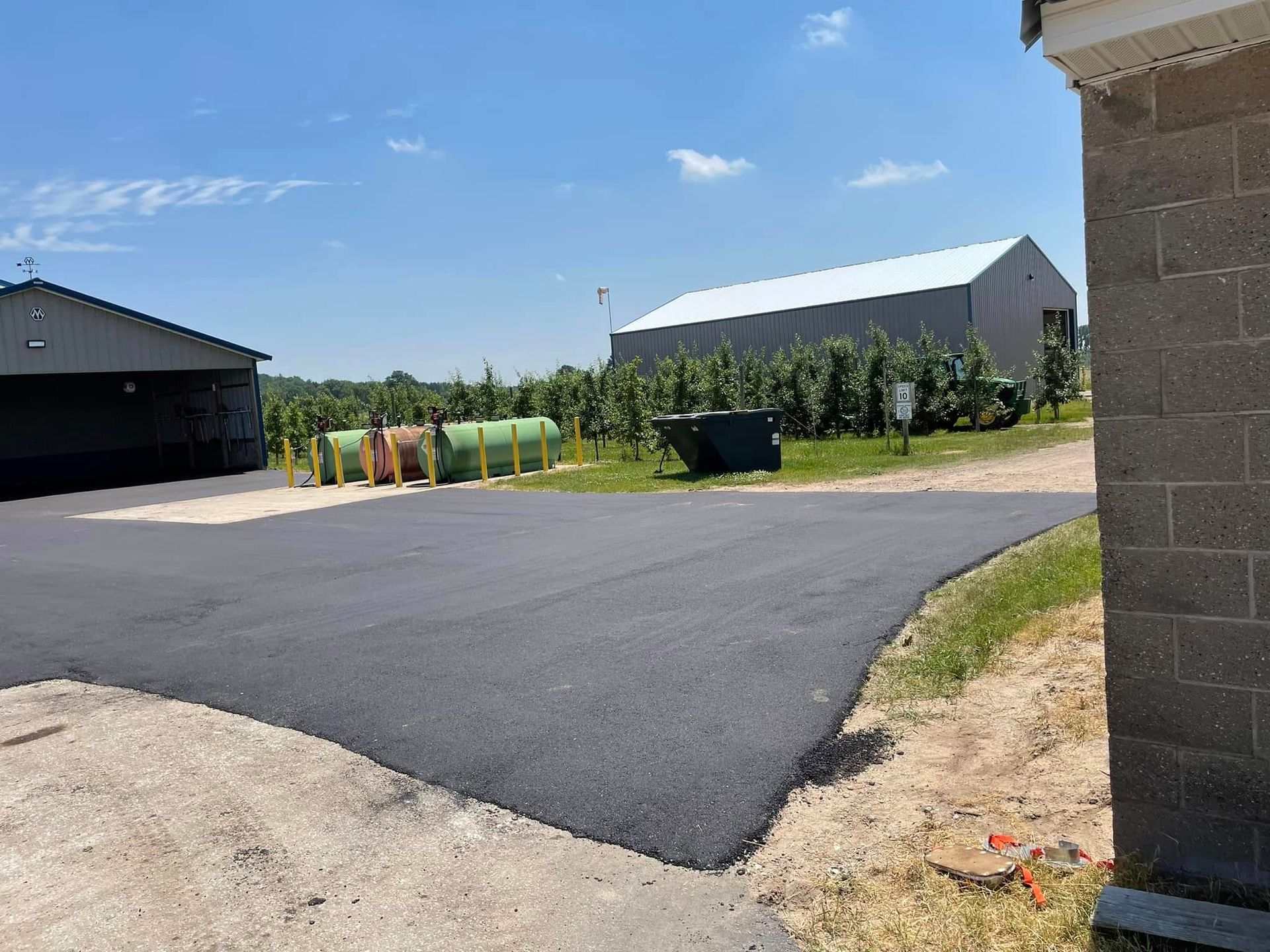 Quiet suburban driveway with fresh black asphalt, a garage, lawn, and a white house under a clear blue sky.