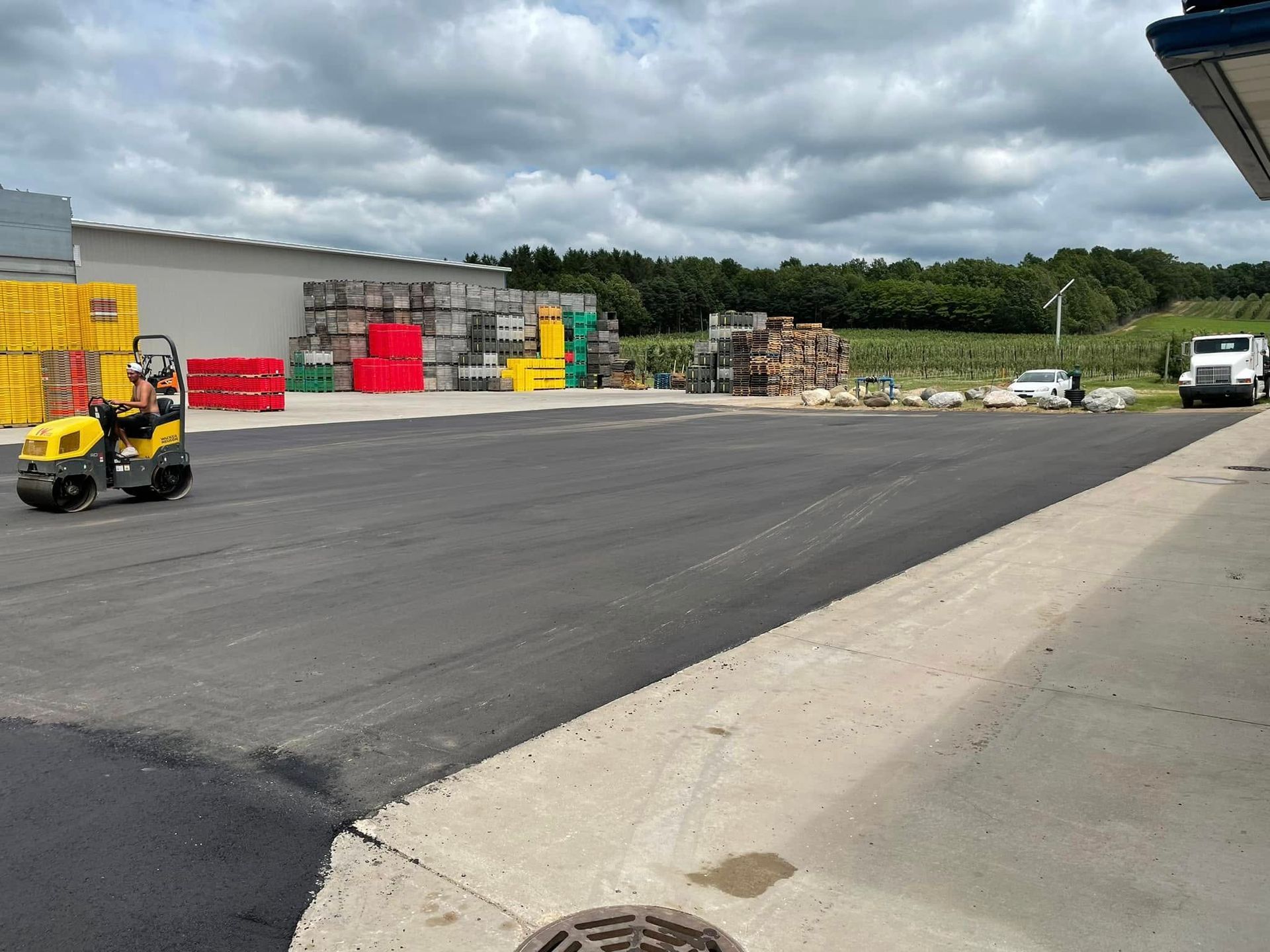 Forklift in a paved industrial yard with stacked pallets, red bins, and a white truck under cloudy skies