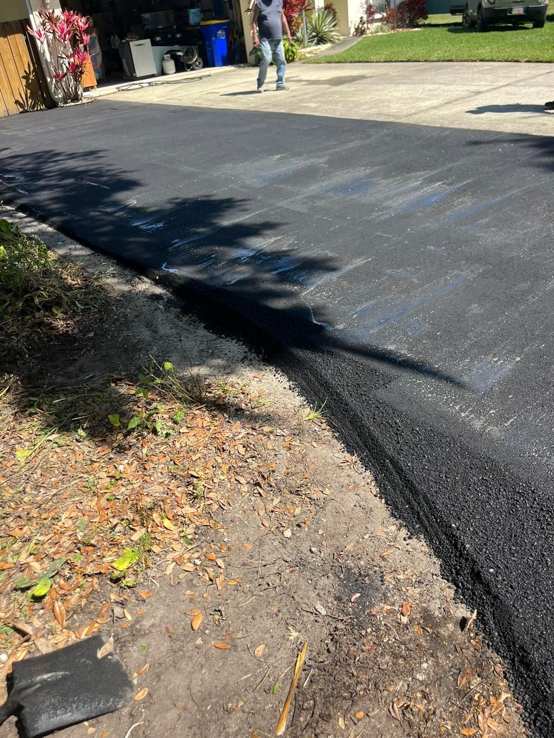 Fresh black asphalt road curving past a dirt shoulder, with a person in the distance near houses and trees