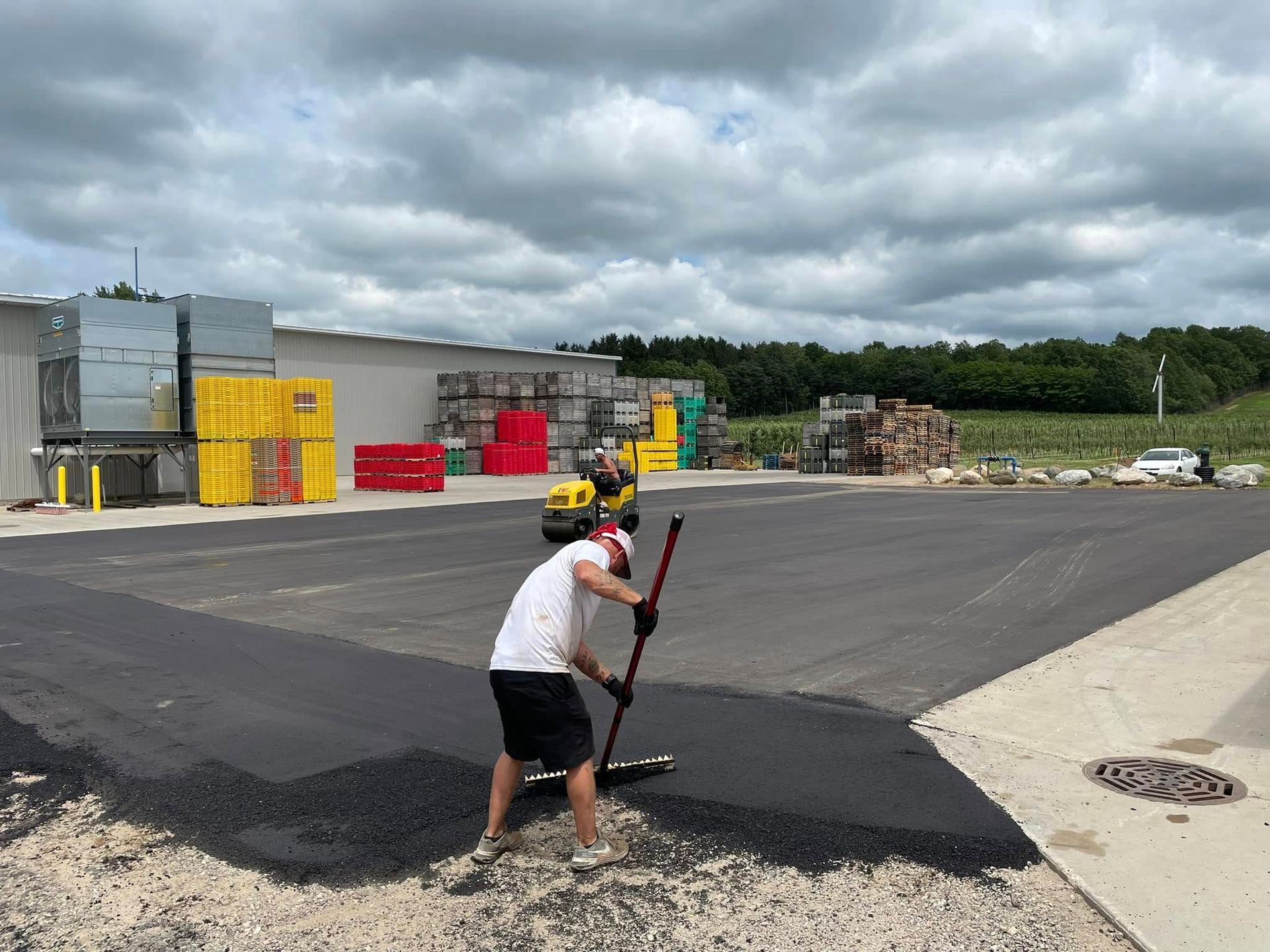 Person shovels black asphalt in a parking lot near industrial buildings under cloudy skies