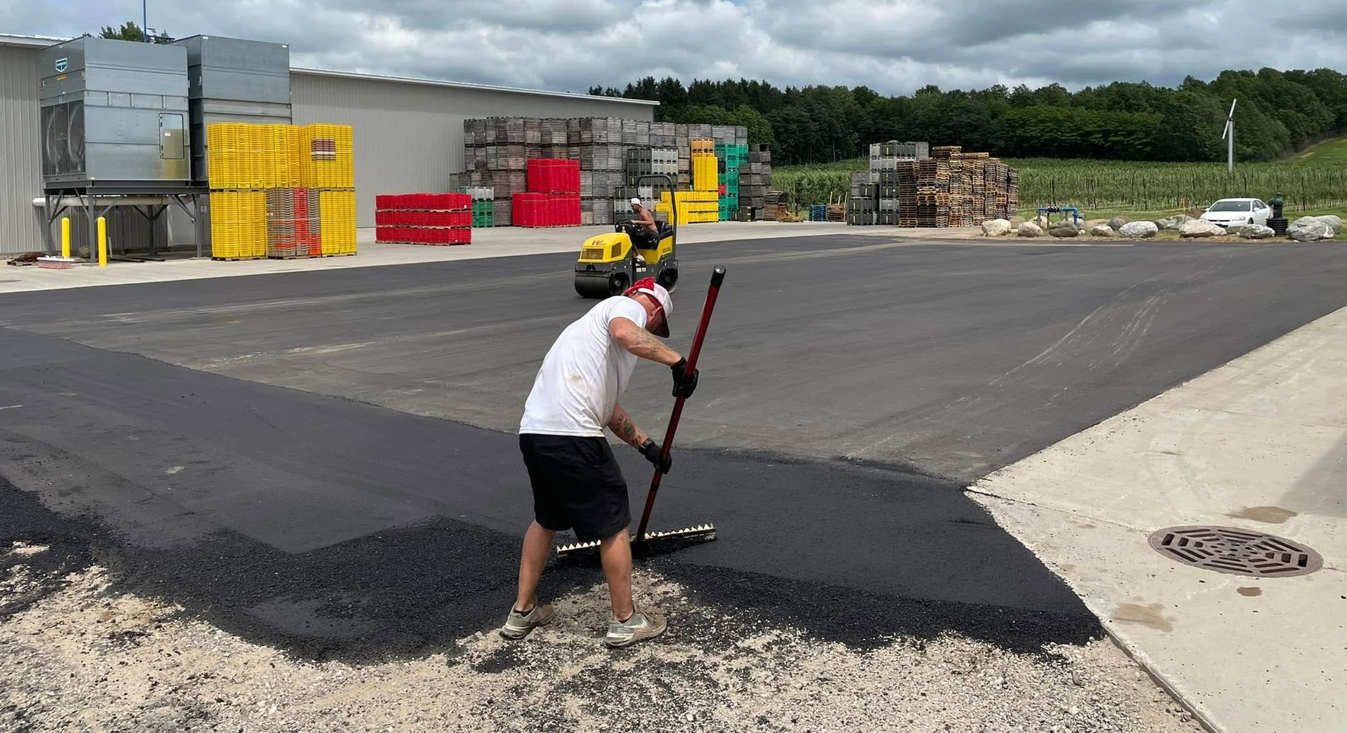 Worker paving a large asphalt lot with a shovel near stacked construction materials.