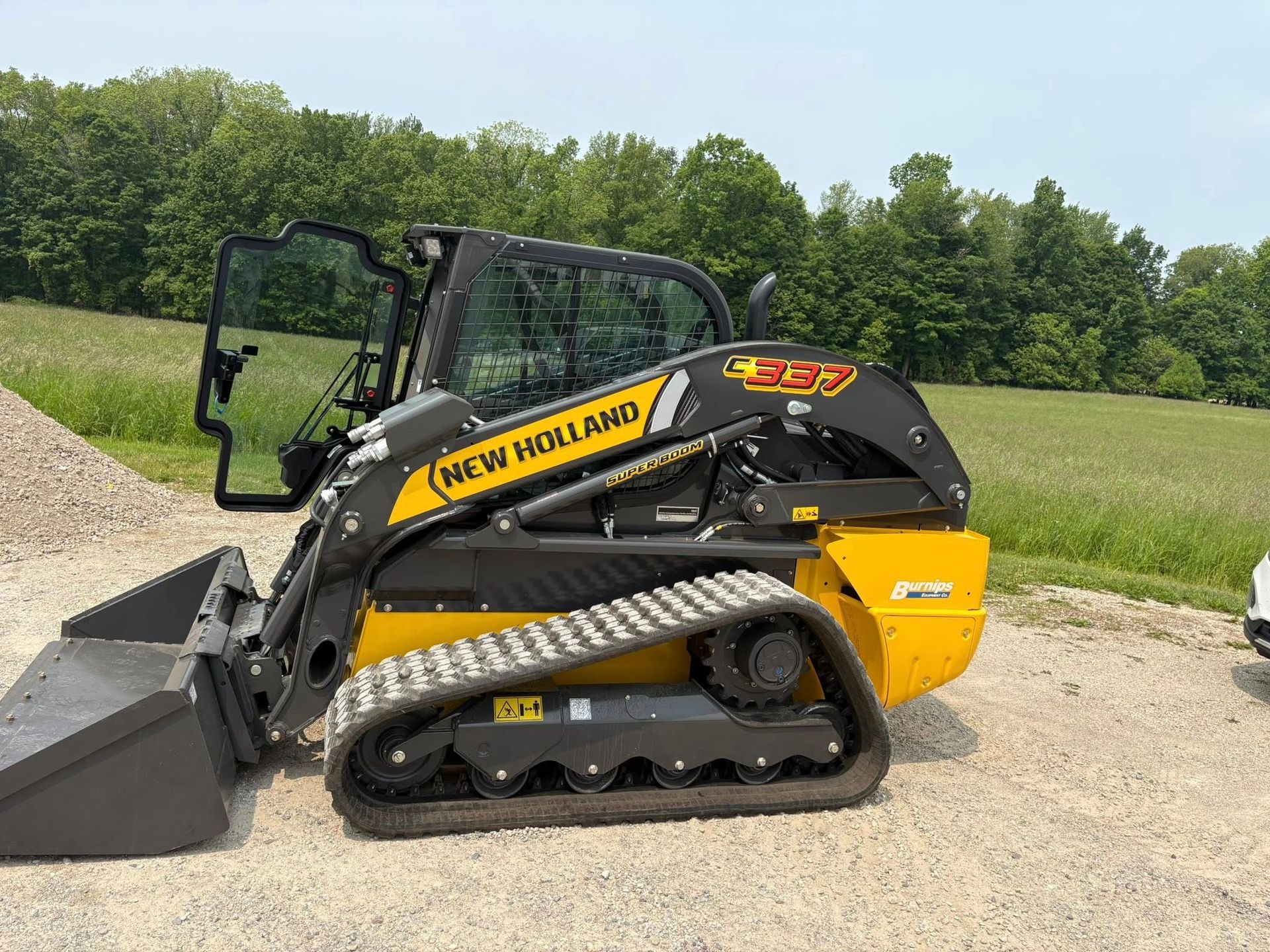 Black-and-yellow compact track loader with bucket, parked on gravel near a grassy field and trees.