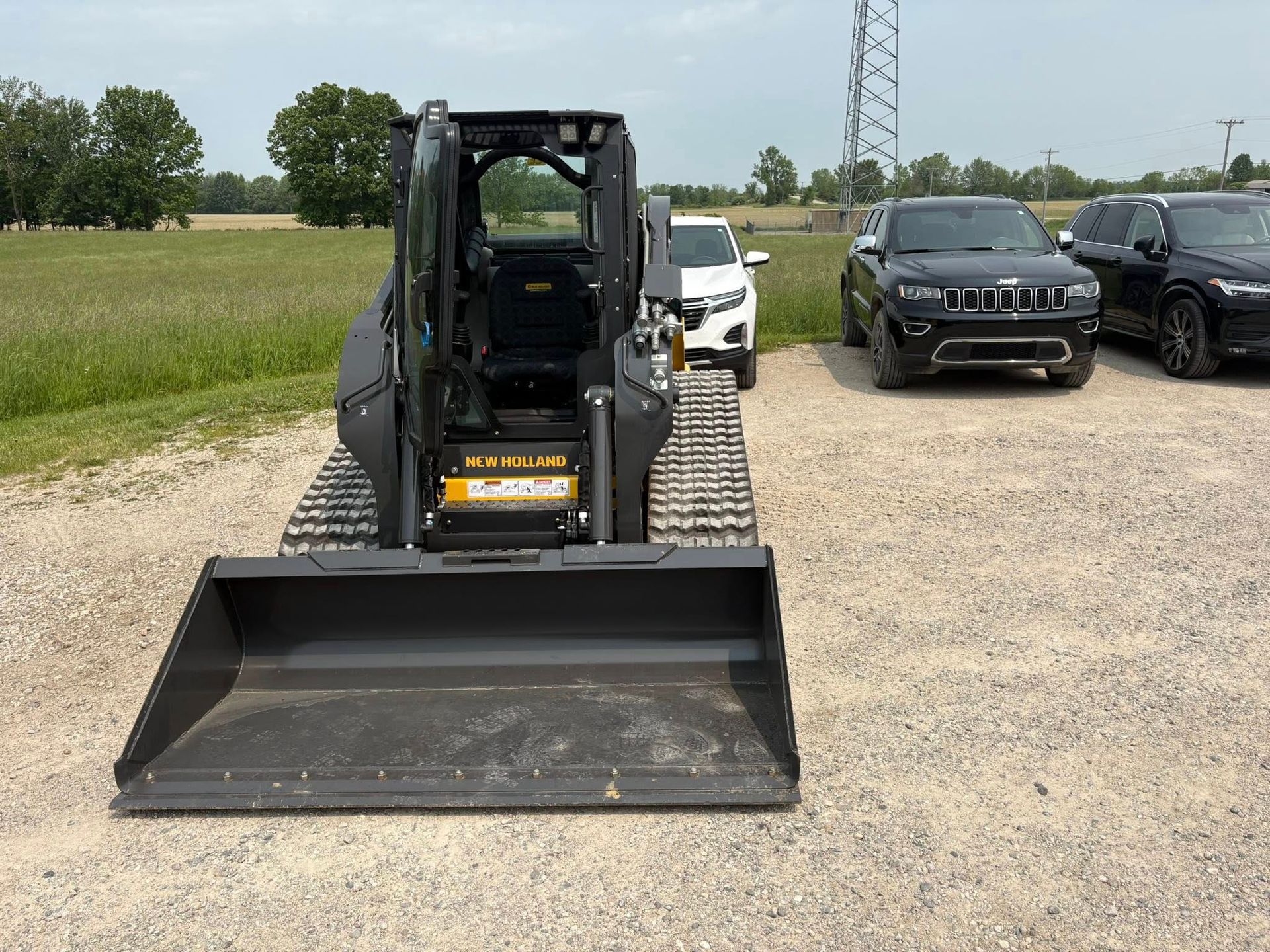 Yellow skid steer with front bucket parked on gravel lot beside cars and open field.