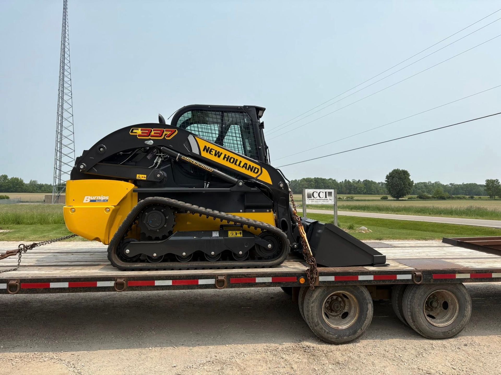 Yellow-and-black skid steer loader on a flatbed trailer in an open field under cloudy skies
